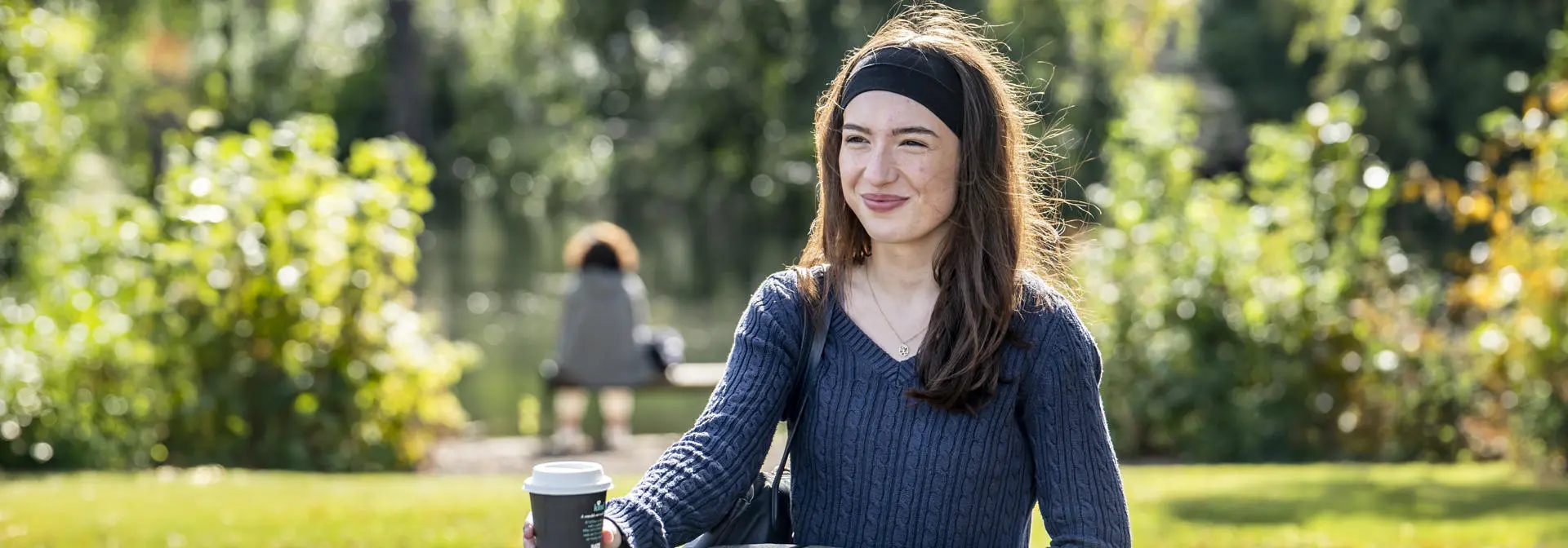 A student sits at a wooden table on campus, holding a coffee cup, surrounded by lush greenery.