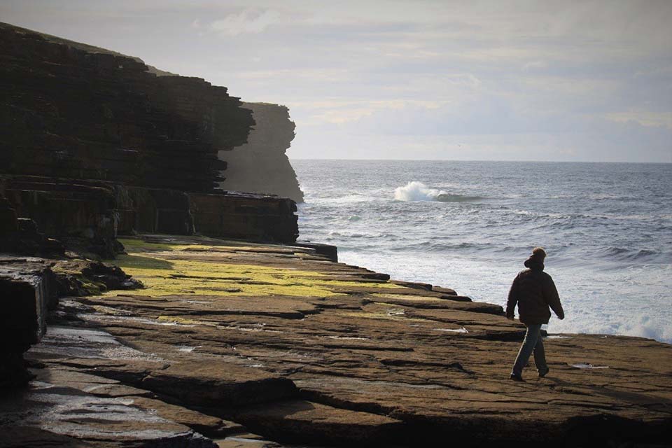 Student walking on Orkney coast as waves crash against cliff face