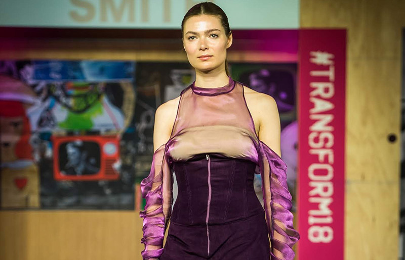 female walking catwalk, wearing an open shoulder iridescent purple top with ruched sleeves and a dark purple corset style high waisted trousers  