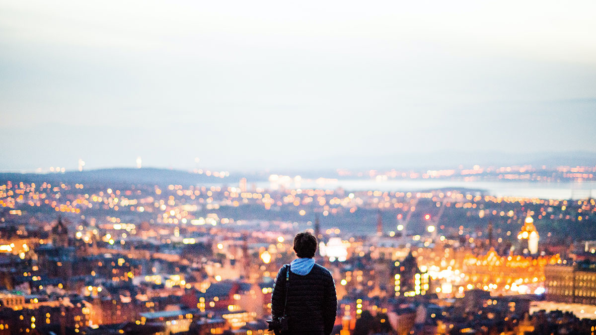 Young person carrying camera looks out across the Edinburgh skyline illuminated at dusk from Arthur's Seat
