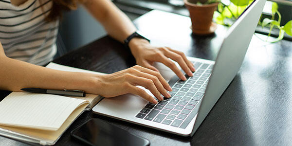 Woman's hands typing on a laptop beside an open notebook