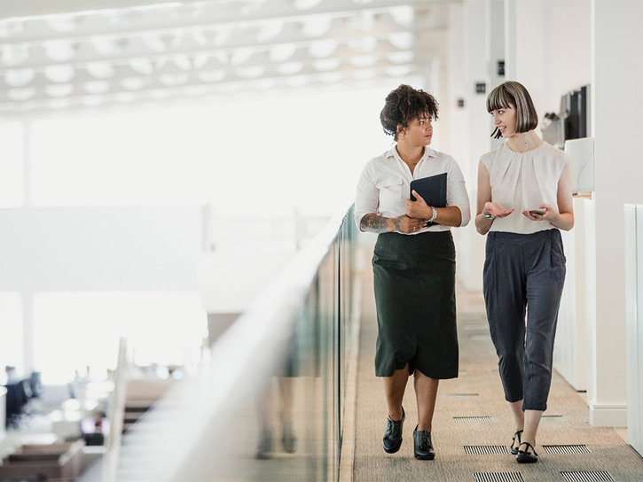 Two female colleagues converse while walking along a mezzanine floor