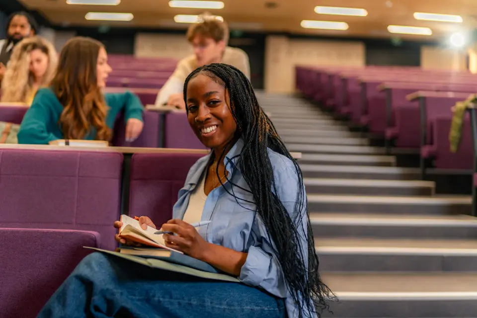 Female student in lecture theatre, making notes and smiling at camera