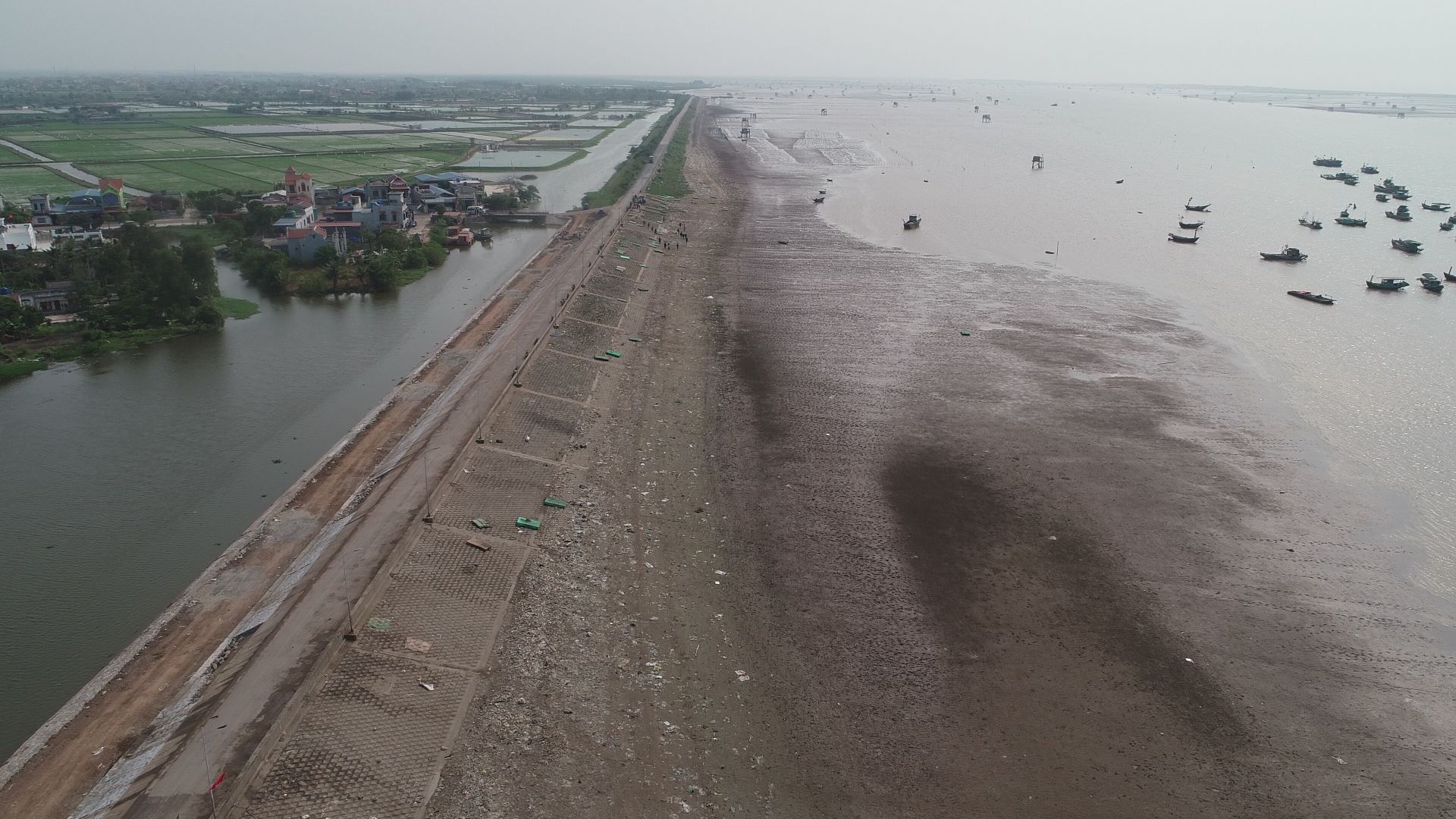 Aerial view of a muddy shoreline with scattered litter, cleanup activity, and boats offshore near a coastal village