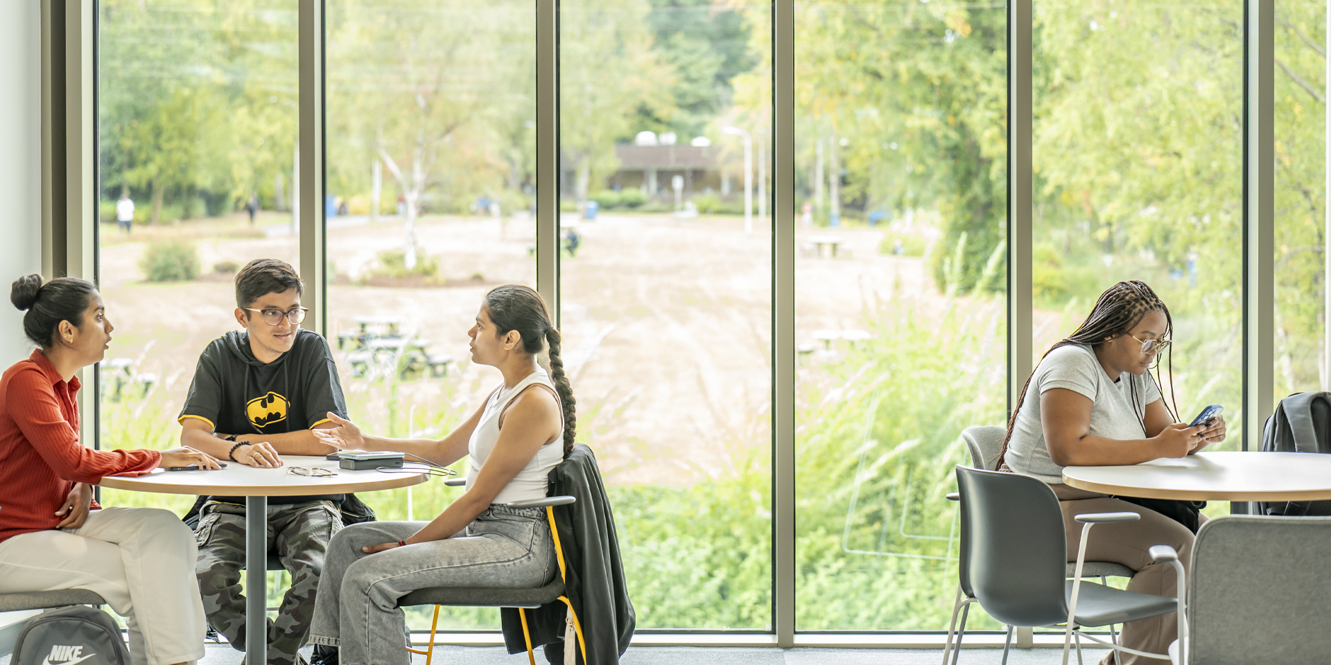 Four students sit at two round tables by large windows, three engaged in conversation and one focused on her phone.
