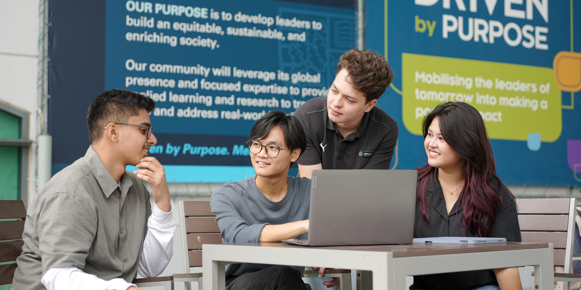 Four students collaboratively working on a laptop at a table, with a colorful wall display behind them.