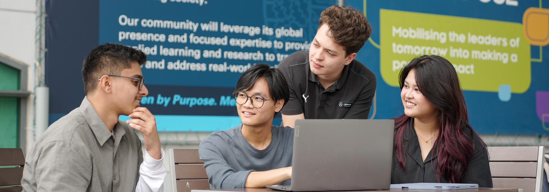 Four students collaboratively working on a laptop at a table, with a colorful wall display behind them.