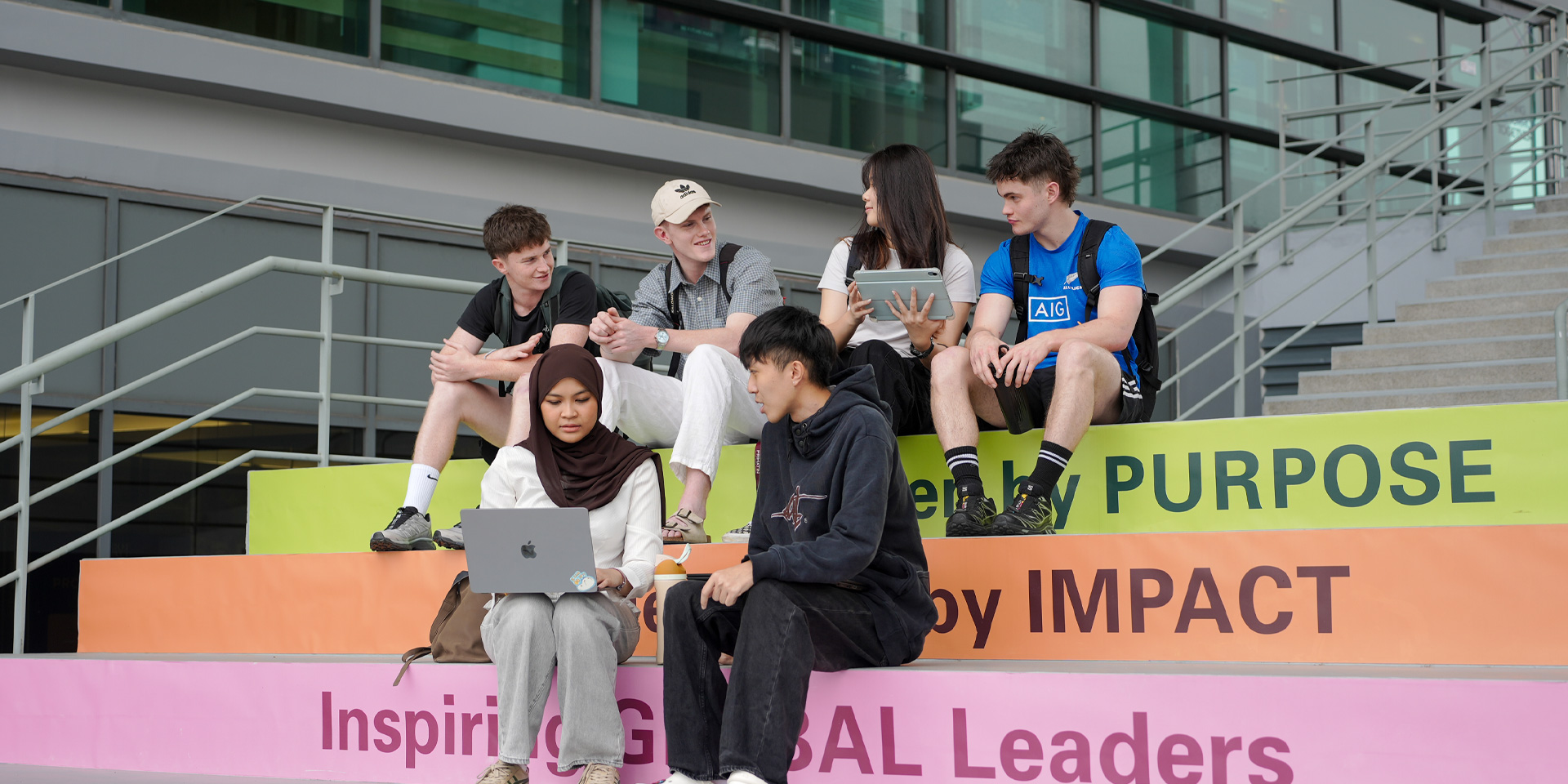Students sit on colorful steps, engaged with laptops and tablets, in a modern outdoor setting.