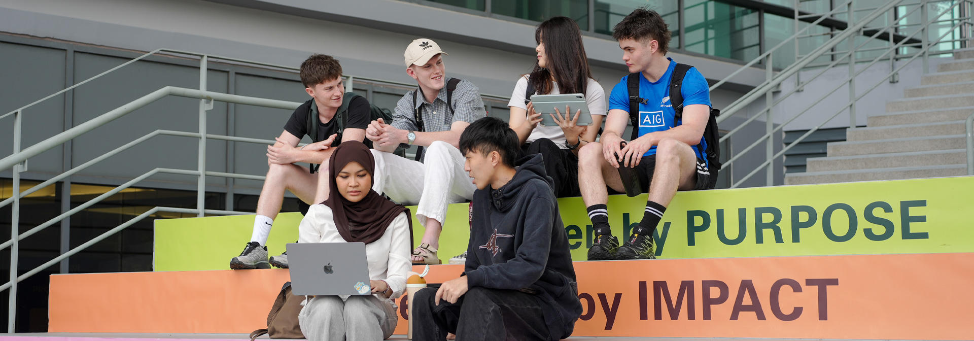 Students sit on colorful steps, engaged with laptops and tablets, in a modern outdoor setting.