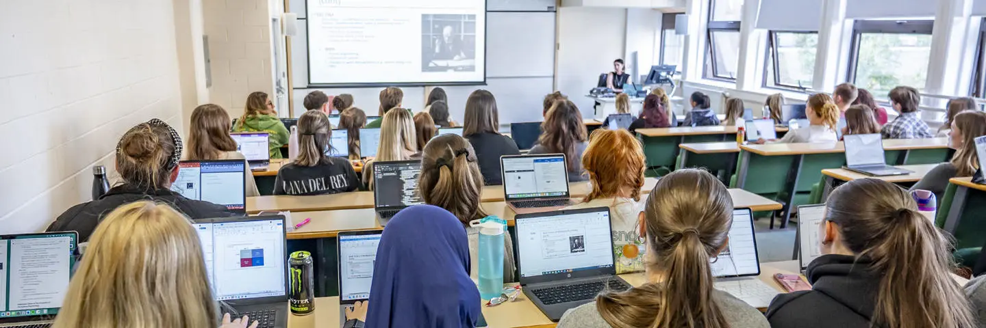 A classroom filled with students using laptops, with a projector displaying content at the front of the room.