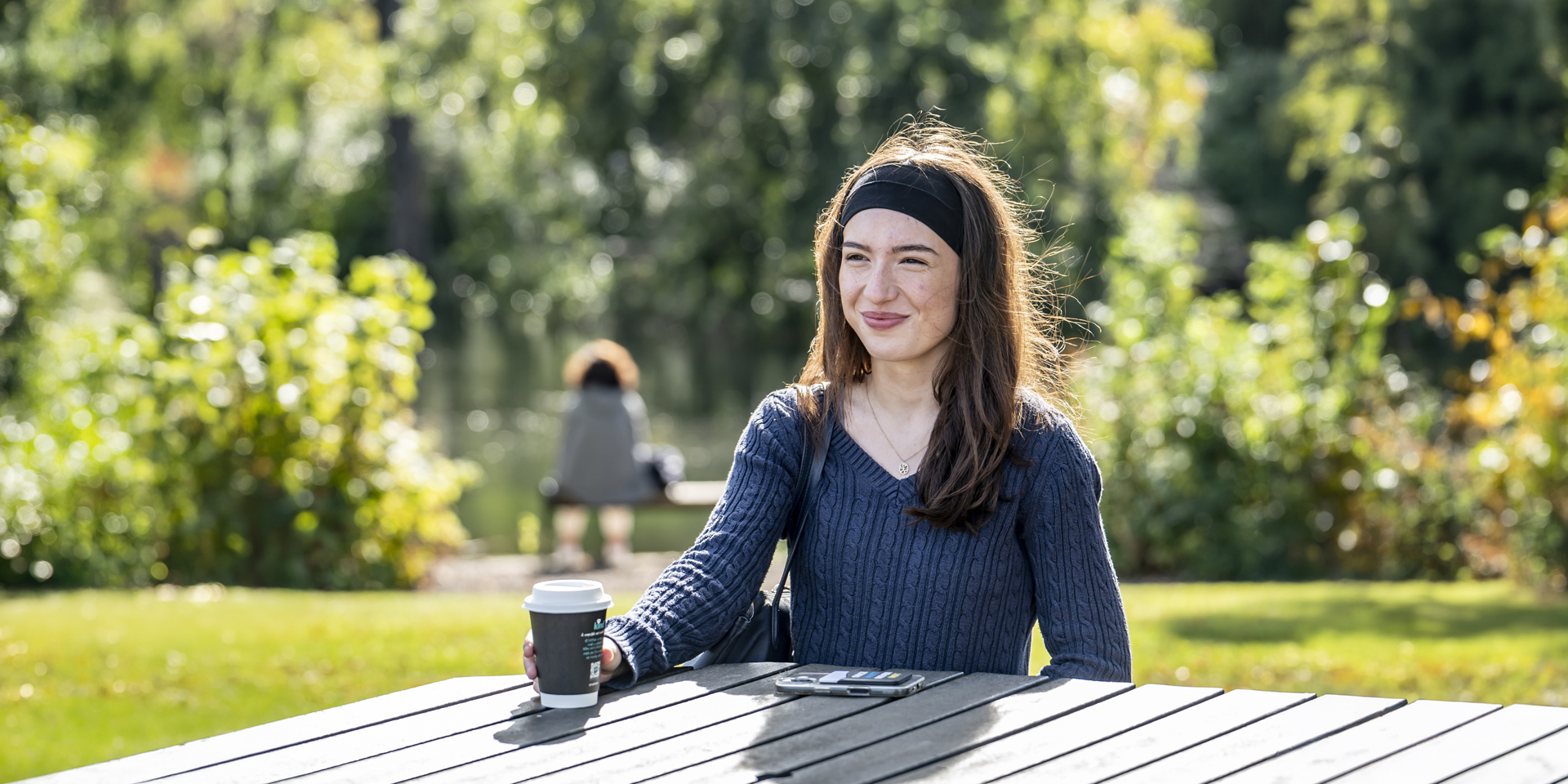 A student sits at a wooden table on campus, holding a coffee cup, surrounded by lush greenery.