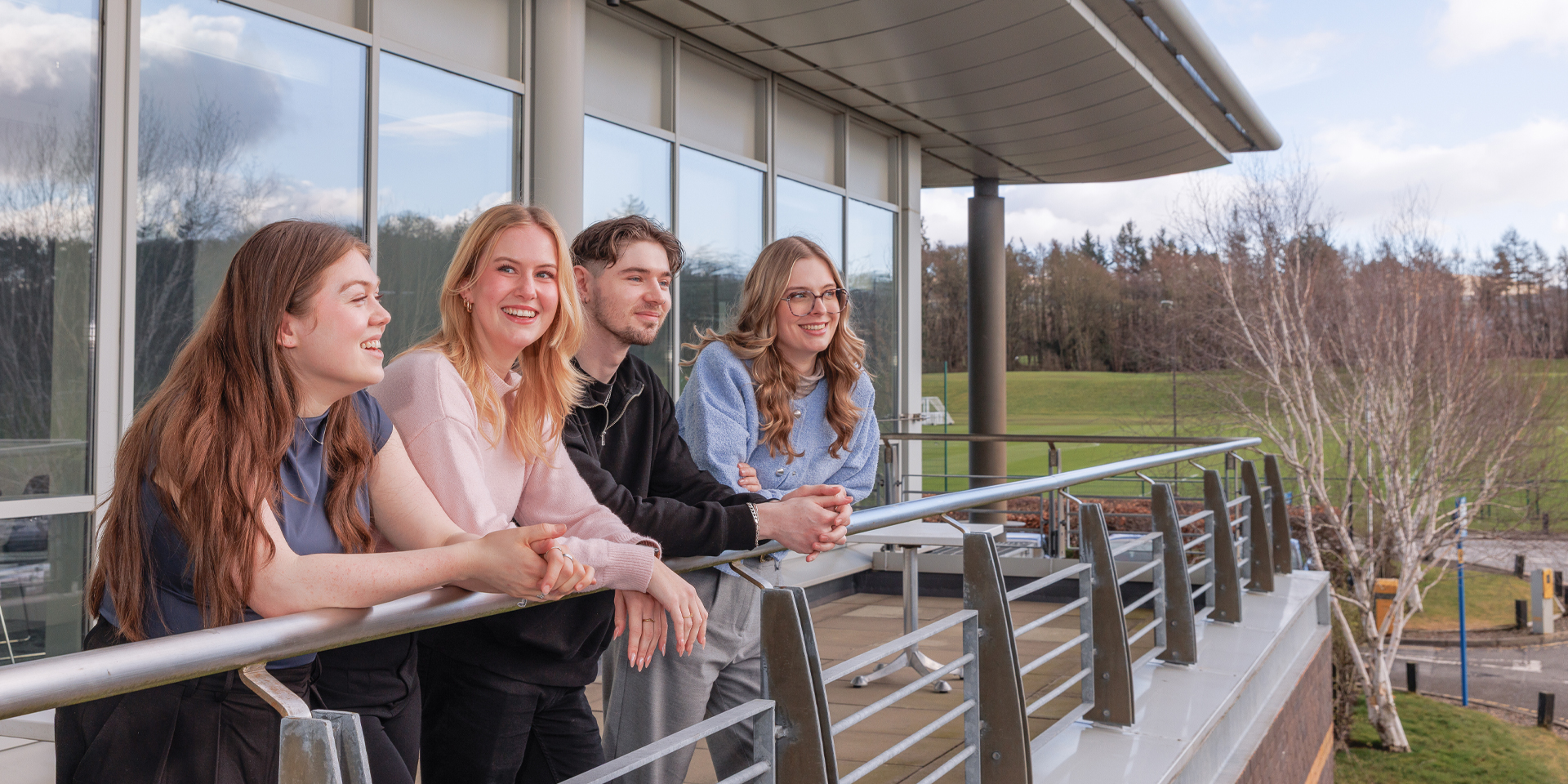 Students lean against a railing on a balcony, enjoying a scenic view of greenery and trees under a partly cloudy sky.