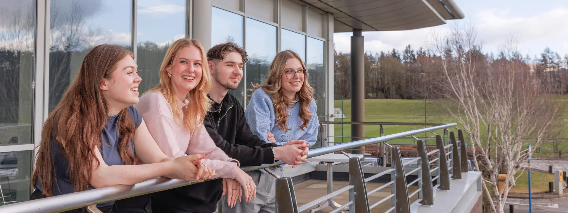 Students lean against a railing on a balcony, enjoying a scenic view of greenery and trees under a partly cloudy sky.