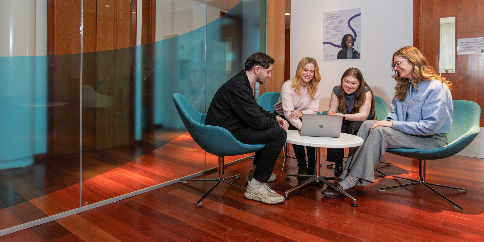 A group of four students collaborates around a laptop in a modern office space.