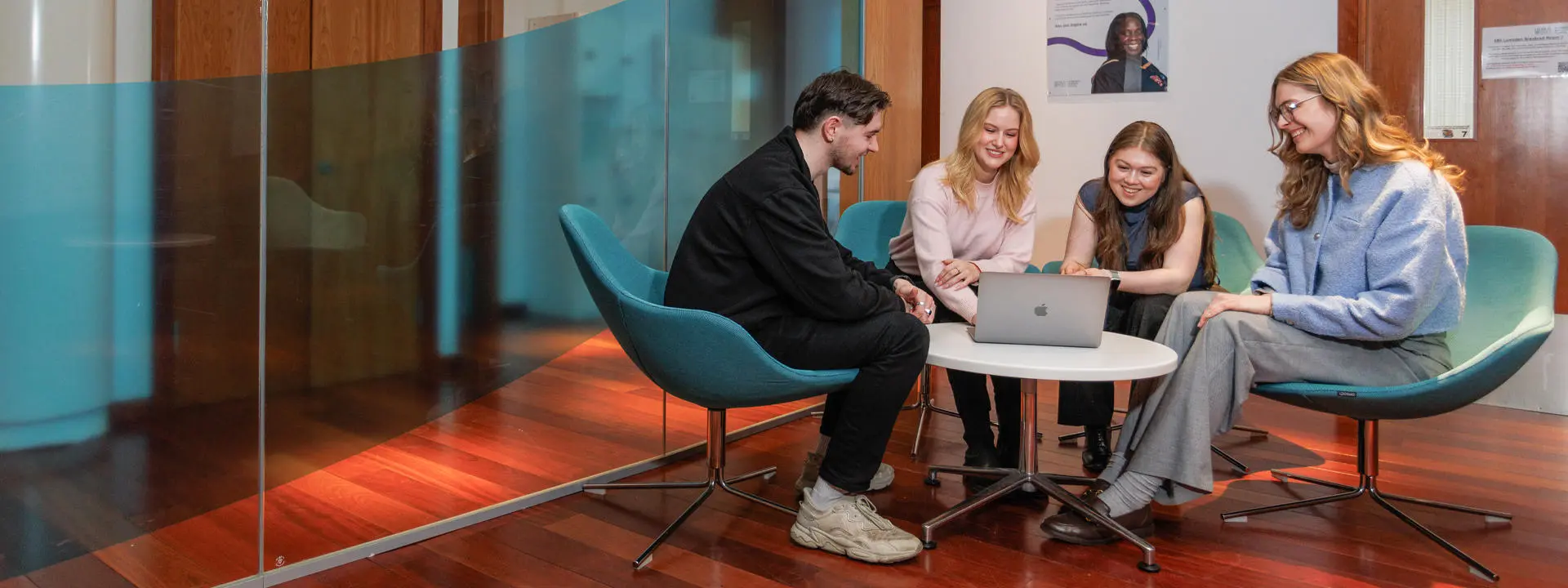A group of four students collaborates around a laptop in a modern office space.
