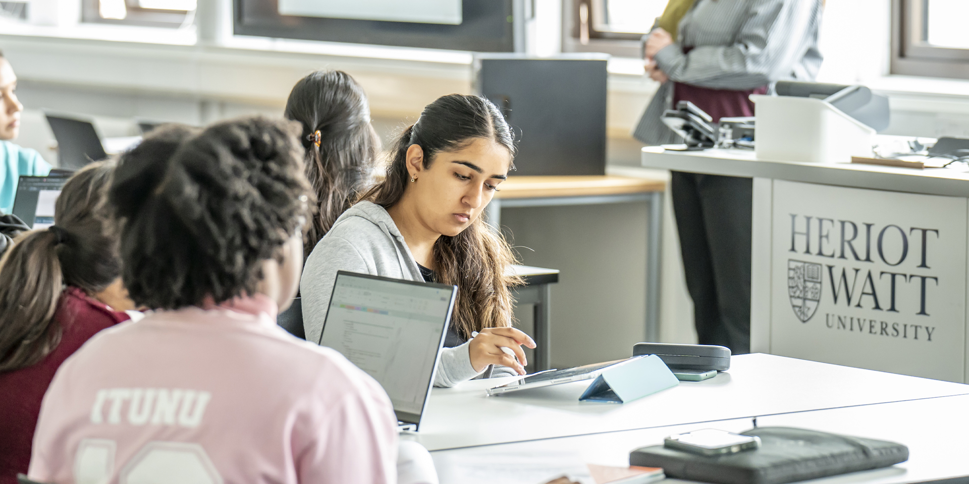Students engaged in a classroom setting, using laptops and tablets with a lecturer in the background.