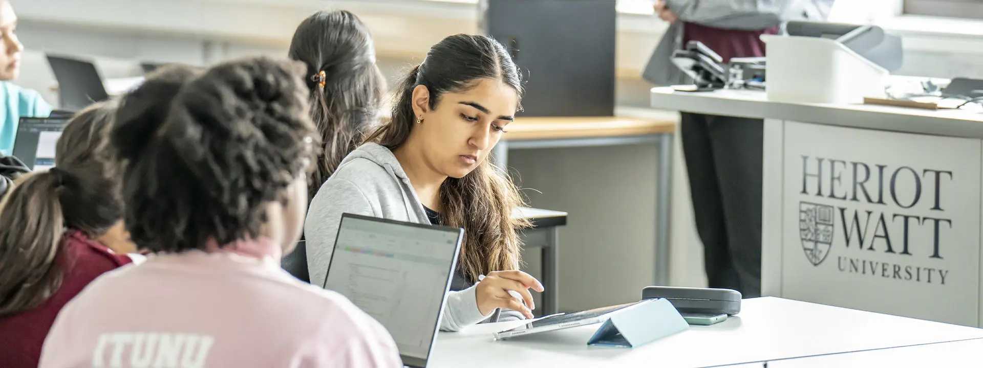 Students engaged in a classroom setting, using laptops and tablets with a lecturer in the background.
