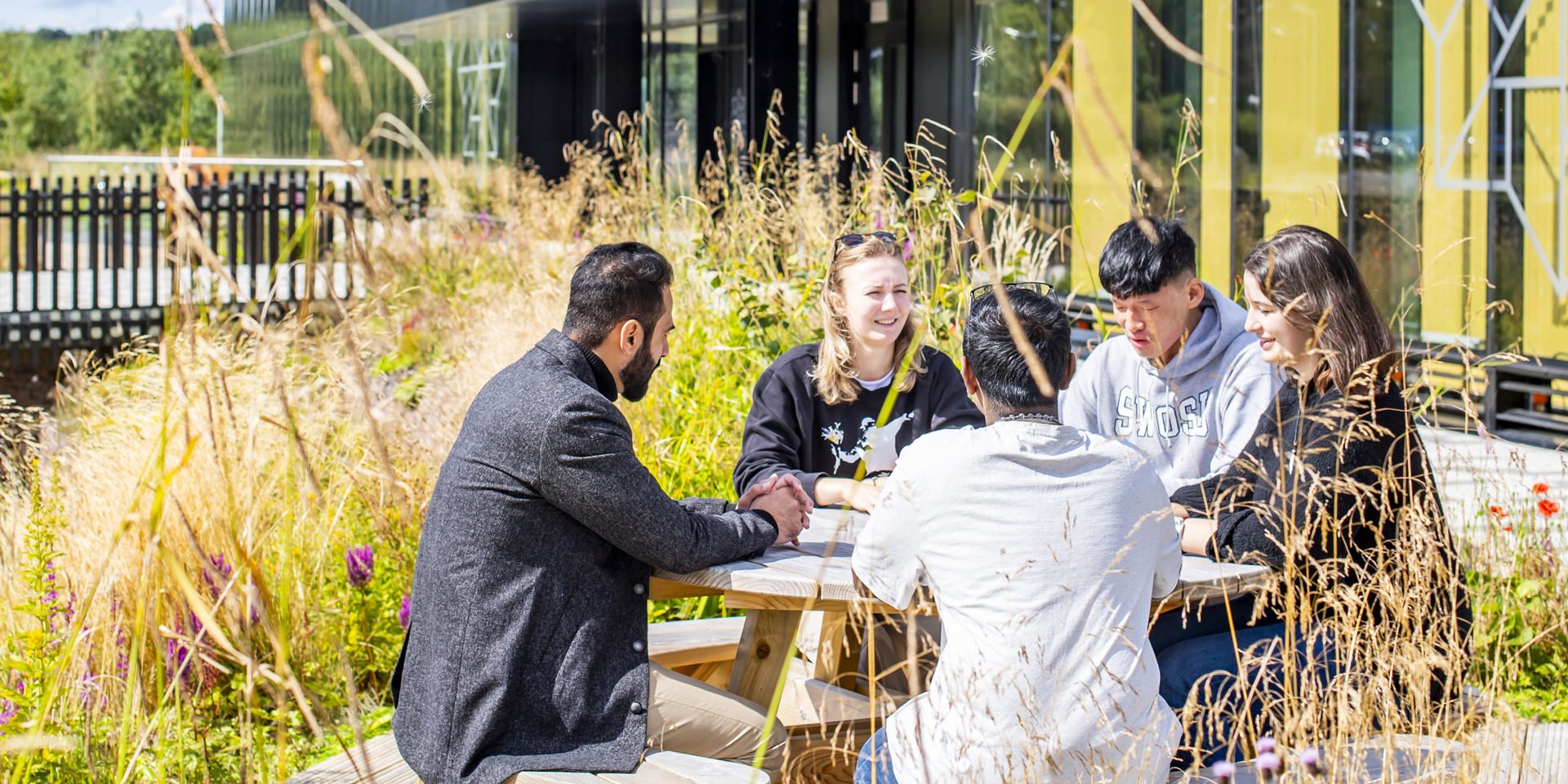 Students sitting around a wooden picnic table outdoors, engaged in conversation.