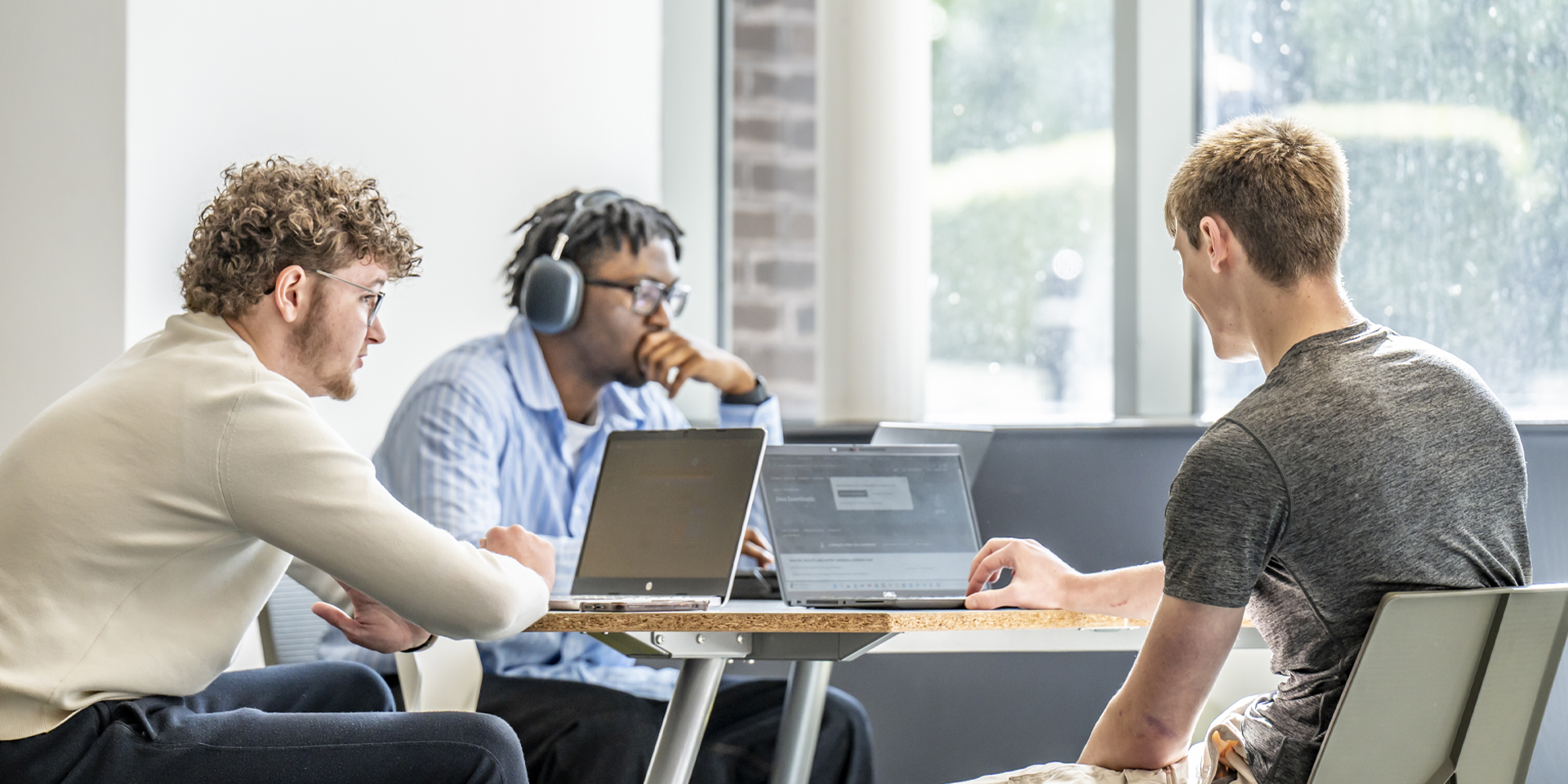 Three students collaborate around a table with laptops, focusing on their screens in a bright, modern study space.