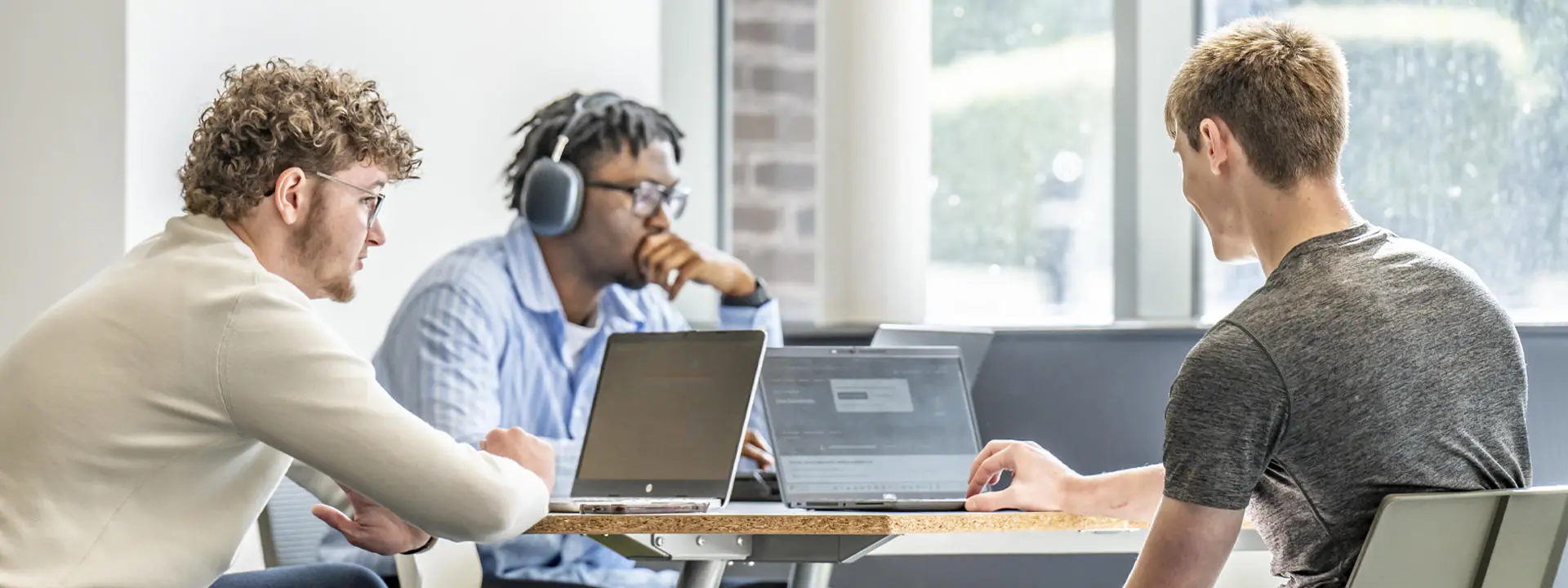 Three students collaborate around a table with laptops, focusing on their screens in a bright, modern study space.
