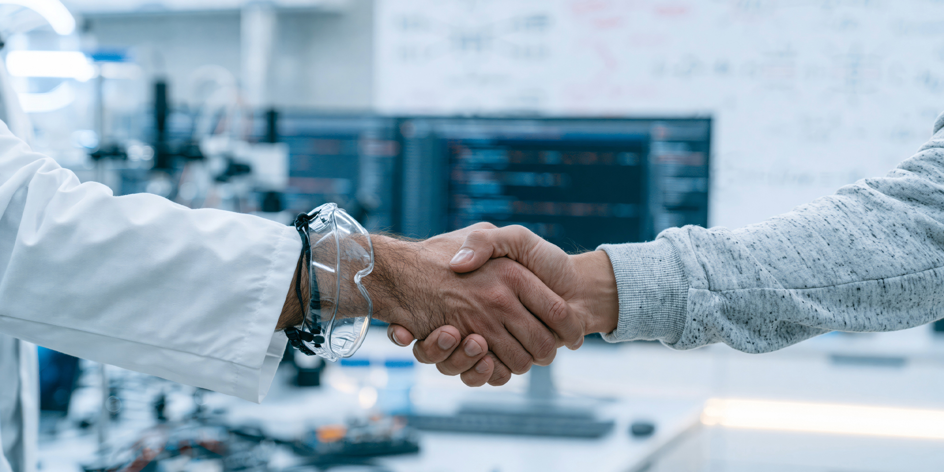 Two hands in a handshake. One with lab goggles and lab coat and another in a jumper. 