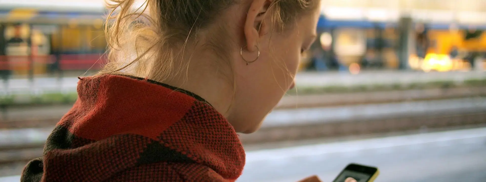 A young woman studies her mobile phone while standing on a train station platform