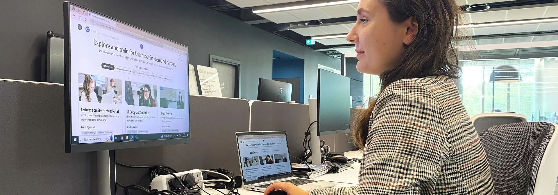 A woman sitting at a desk in an office browsing online courses on a computer.