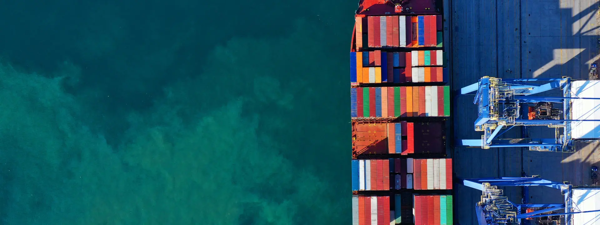 Aerial view of a cargo ship docked at a port, loaded with colourful shipping containers beside blue cranes.
