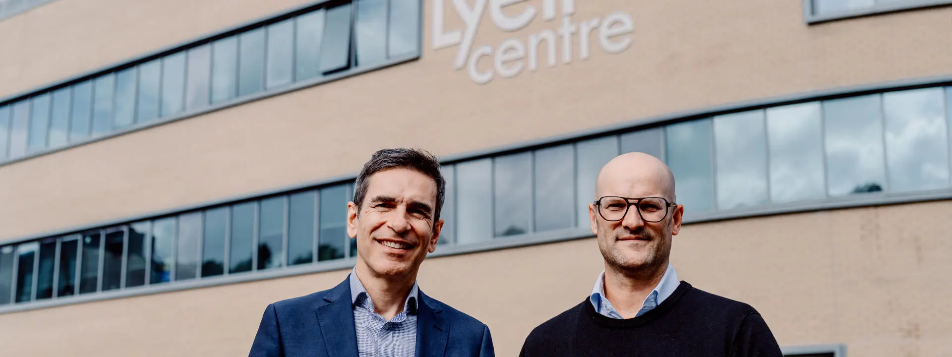 Two men stand in front of the Lyell Centre building. It is a large brick building with lots of windows.