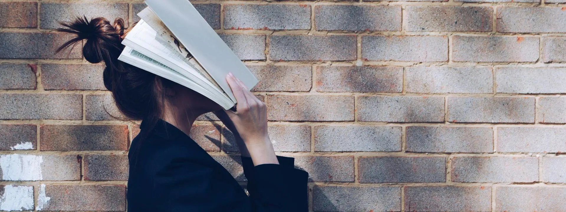 Girl in front of red brick wall holding book up to her face.
