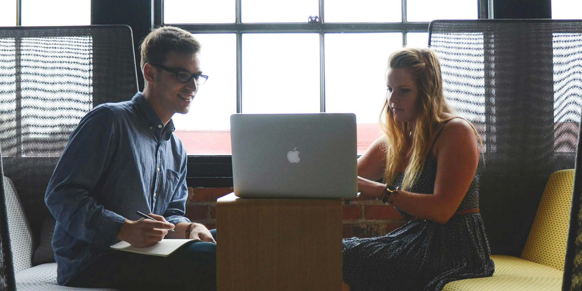 A man and woman are seated at a table, engaged with a laptop in front of them, discussing or collaborating on a project.