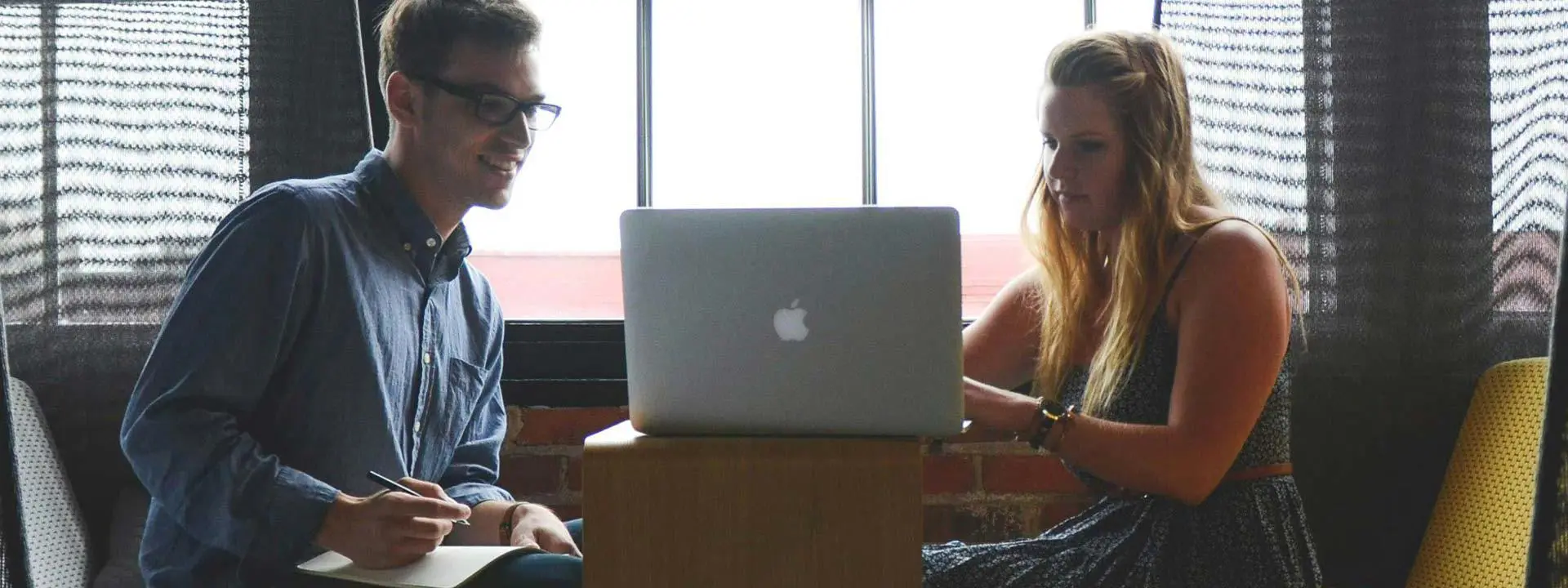 A man and woman are seated at a table, engaged with a laptop in front of them, discussing or collaborating on a project.