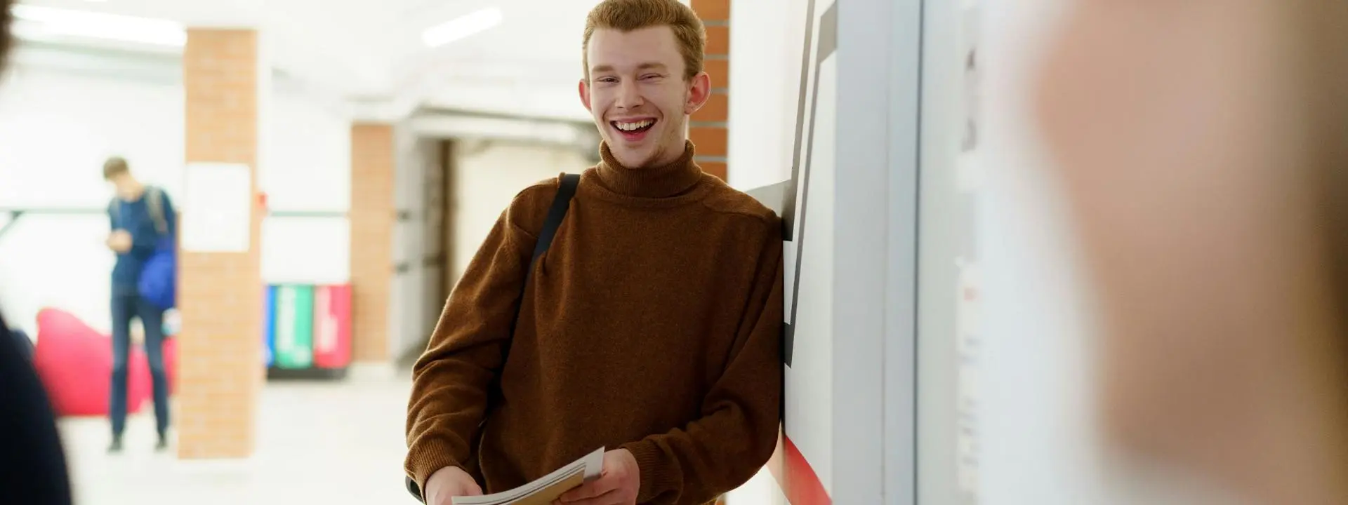 A young man wearing a brown polo neck jumper leans against the wall and laughs