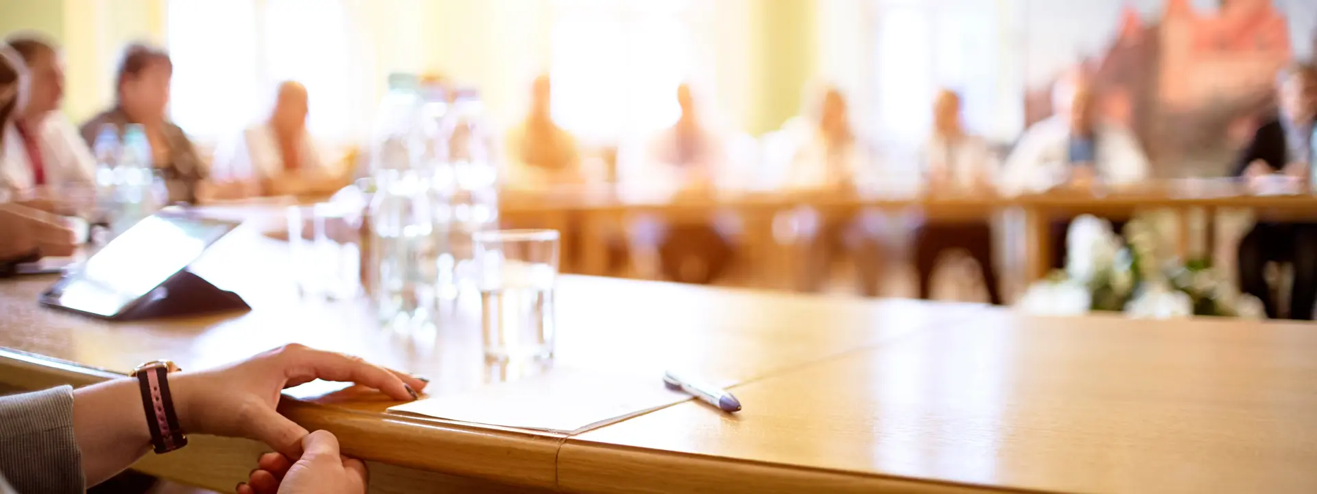 Person seated at a conference table, with a group discussion taking place in the background in a meeting room.