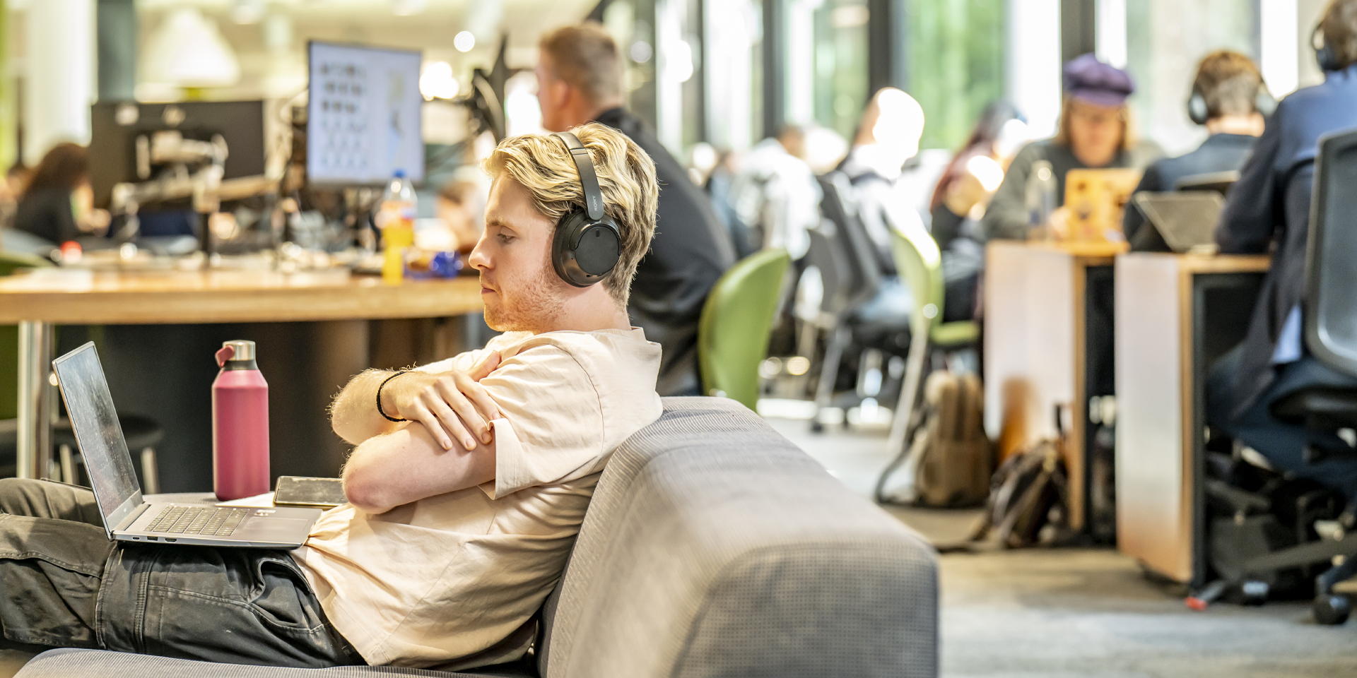 A student seated on a couch, wearing headphones, using a laptop in a busy environment.