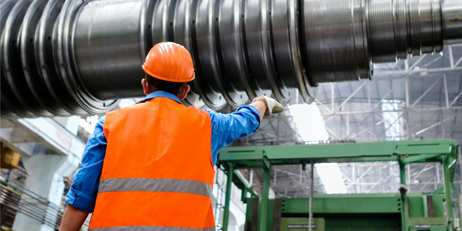 Worker in orange safety vest and helmet points at large industrial machinery inside factory.