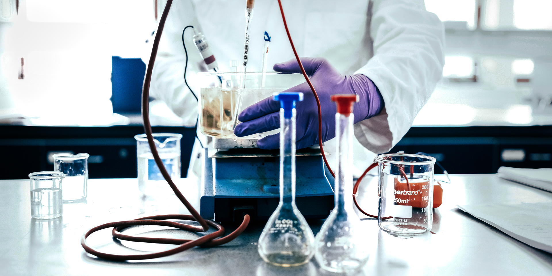 Student in purple gloves working with beakers and electrodes on a lab bench with scientific instruments and wires.