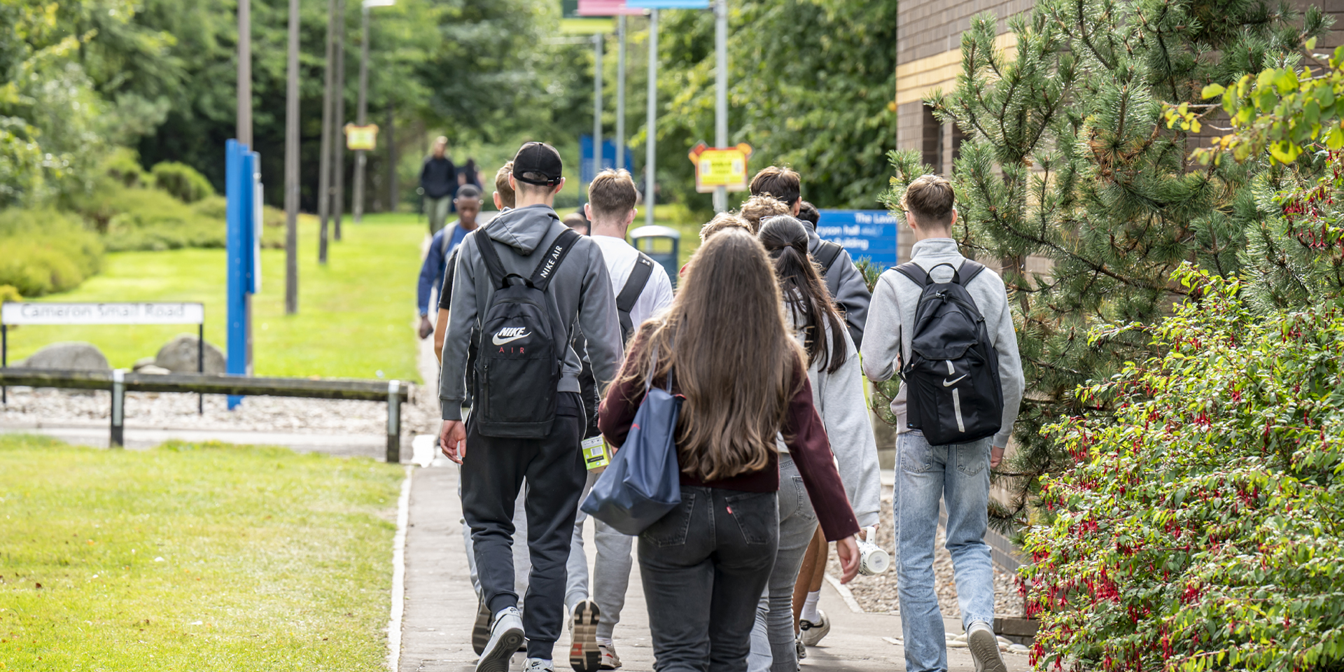 Students walking down a path lined with greenery, heading towards a building with signs and flags in the background.