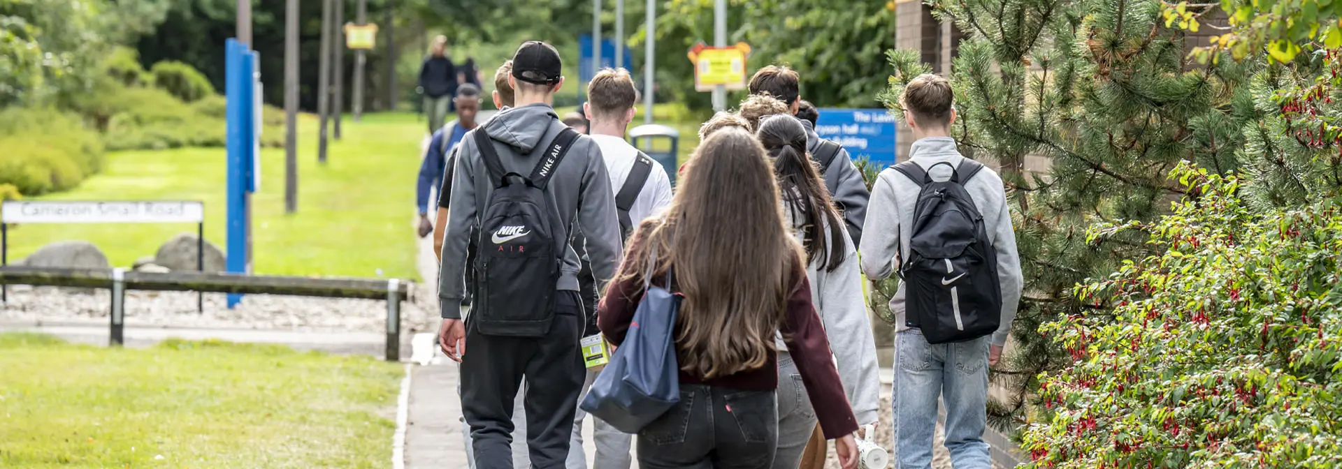 Students walking down a path lined with greenery, heading towards a building with signs and flags in the background.