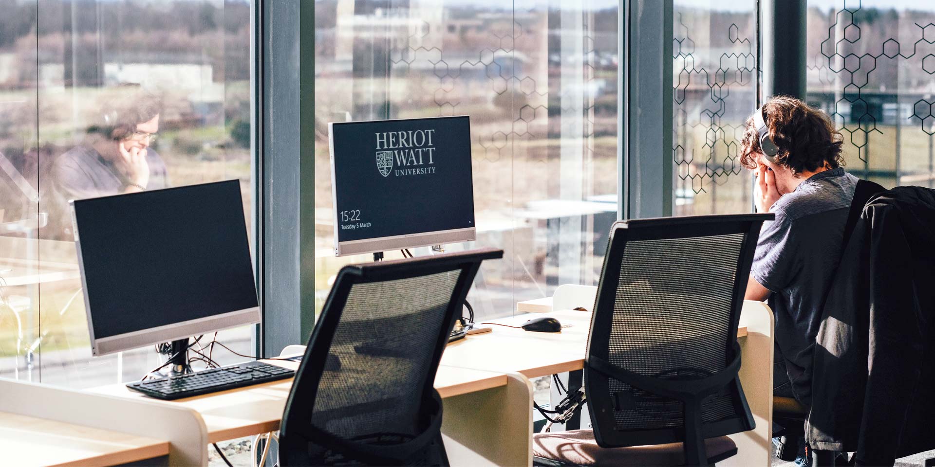A student seated at a desk, working on two computers, focused on his tasks.