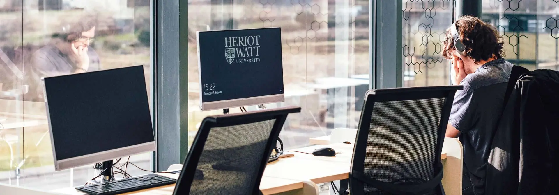 A student seated at a desk, working on two computers, focused on his tasks.