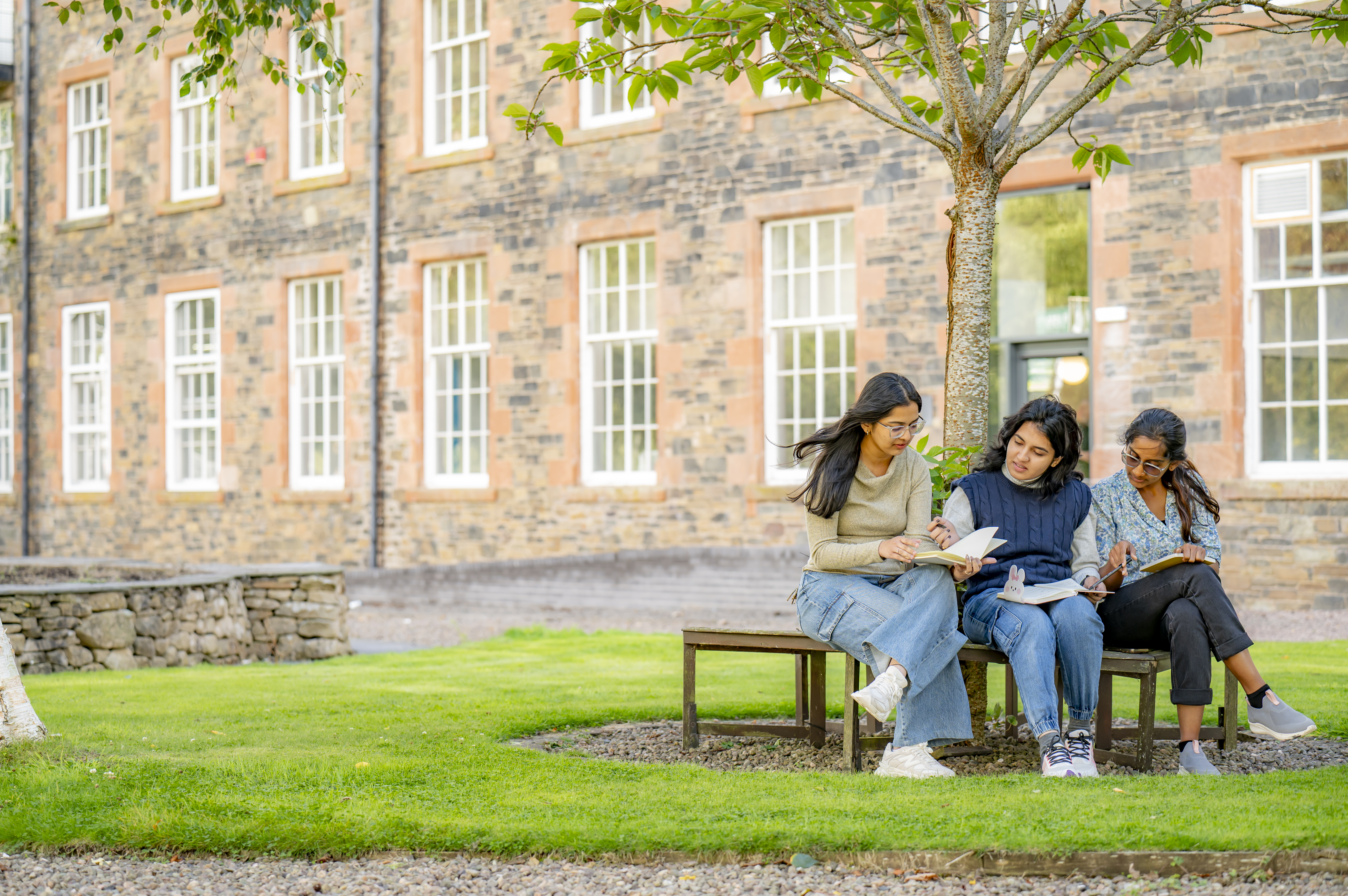 Three students sat on a bench underneath a tree on green grass, outside Heriot-Watt's Scottish Borders Campus building