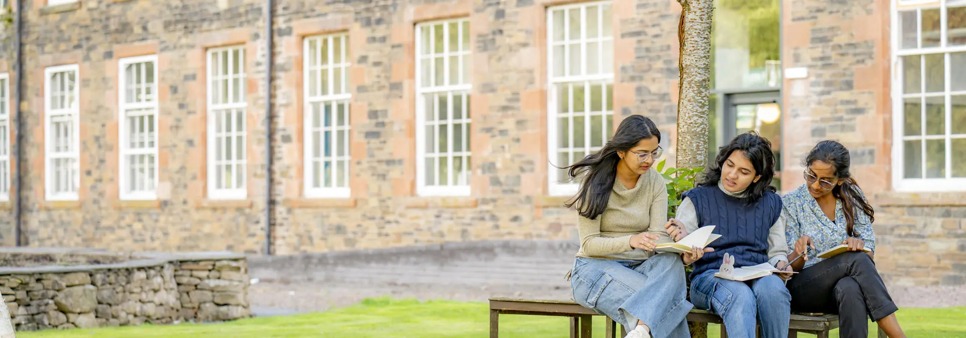 Three students sat on a bench underneath a tree on green grass, outside Heriot-Watt's Scottish Borders Campus building
