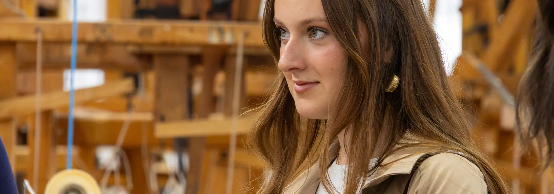 Young woman wearing a beige coat looking to the side, with a lot of wooden weaving machinery in the background.