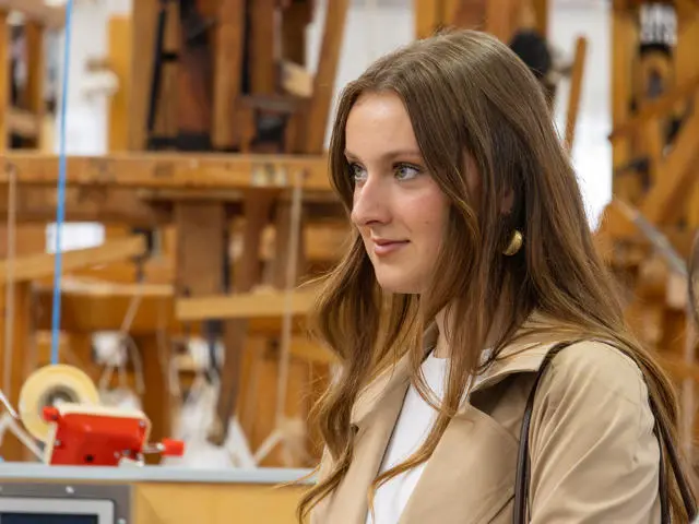 Young woman wearing a beige coat looking to the side, with a lot of wooden weaving machinery in the background.