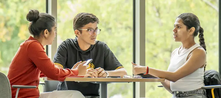Three students engaged in discussion while seated at a table.