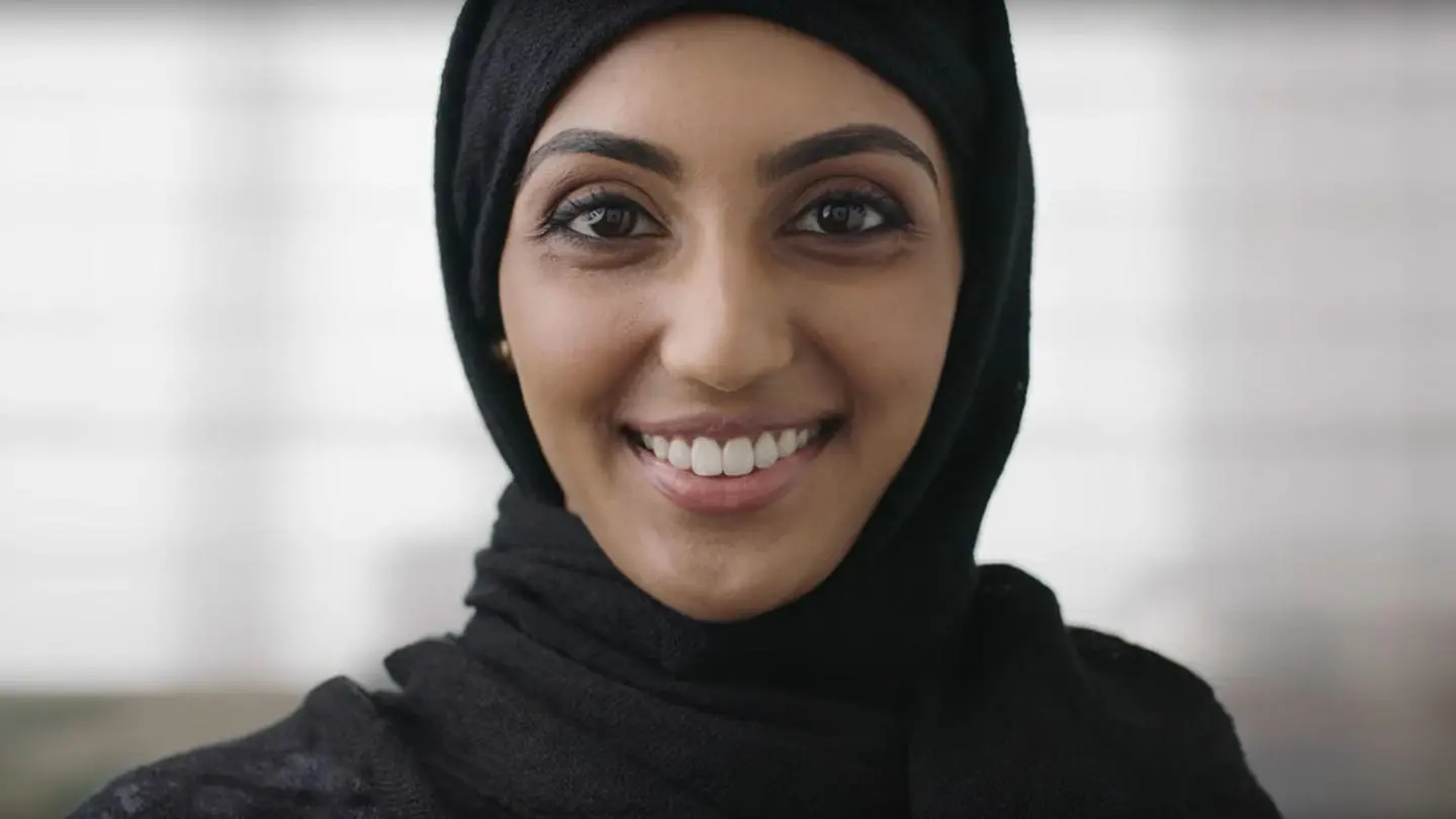 A woman in a black headscarf smiles warmly at the camera, exuding joy and confidence.