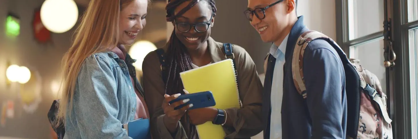 Three students standing in a corridor looking at a phone.