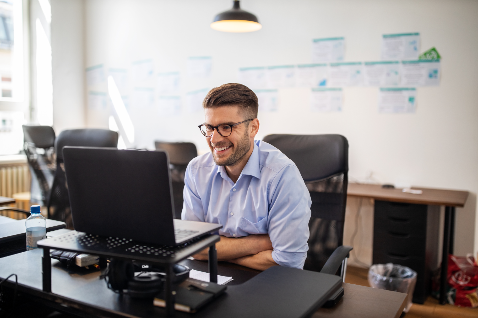 A man wearing glasses smiles as he sits working on a laptop.