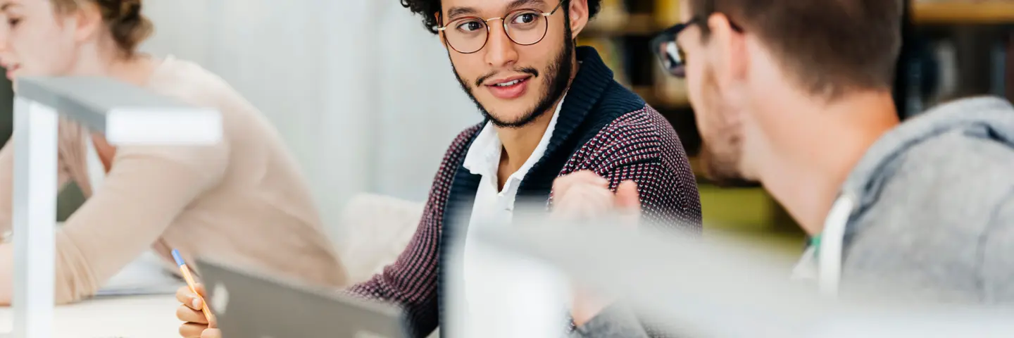 Two men wearing glasses in discussion while working on laptops in a library.